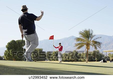 Stock Image - Man playing golf, woman holding golf flag. Fotosearch