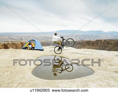 Man riding mountain bike near puddle, woman in tent in background View Large Photo Image Stock Image - Man riding mountain bike near puddle, woman in tent in background. Fotosearch