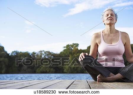 Stock Image - Mature woman sitting crossed legged by lake, eyes closed. Fotosearch
