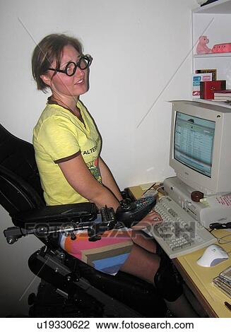 Stock Image - Young woman in glasses uses an elevating wheelchair to access her computer workstation.. Fotosearch