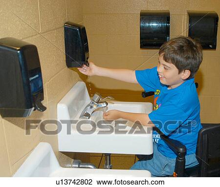 Stock Image - Boy with Muscular Dystrophy, utilizing a scooter/cart for mobility, washing his hands in the restroom.. Fotosearch
