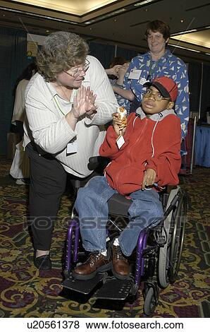 Stock Photo - Little boy with a disability, utilizing a wheelchair for mobility, interacting with aides and friends at a conference. Fotosearch