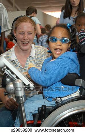 Stock Photo - Little girl with a disability - utilizing a wheelchair for mobility and a communication device for expressive language - enjoying some time in the community with a care provider.. Fotosearch
