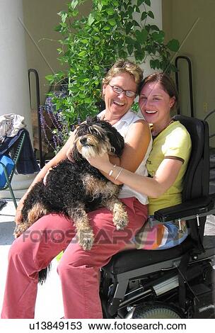 Stock Image - Mother with dog in her lap sits on the lap of her daughter (wheelchair user).. Fotosearch