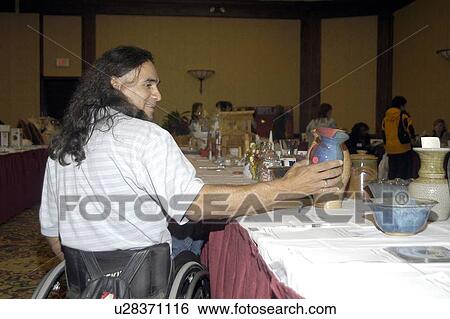 Stock Photograph - Man with a disability, utilizing a wheelchair for mobility, examining items at a silent auction.. Fotosearch