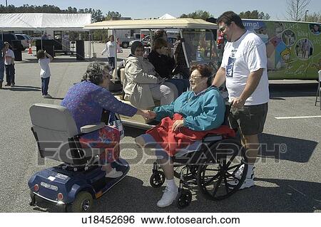 Stock Photograph - Two women with disabilities (a scooter user & a wheelchair user) and a man meet in front of a community fair. Fotosearch