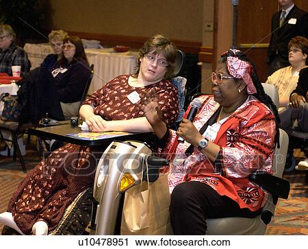 Stock Image - Two women with disabilities, both utilizing wheelchairs for mobility, talking and reflecting about a presentation at a conference for people with disabilities.. Fotosearch