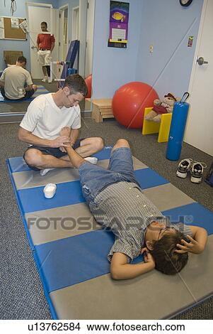 Boy with muscular dystrophy lays on mat as therapist stretches his left leg. View Large Photo Image Picture - Boy with muscular dystrophy lays on mat as therapist stretches his left leg.. Fotosearch