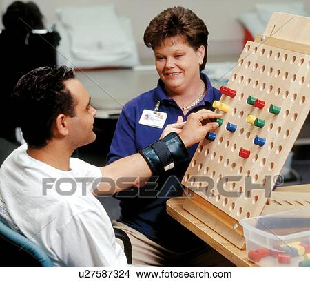 Picture - Man with a disability putting pegs in a board during a session with his occupational therapist.. Fotosearch