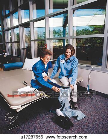 Stock Photograph - Older woman with disabilities receiving assistance in dressing from one of her therapists at a rehabilitation facility.. Fotosearch