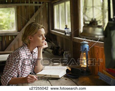 Stock Image - Woman sitting at a table in cabin with book and eyeglasses.. Fotosearch