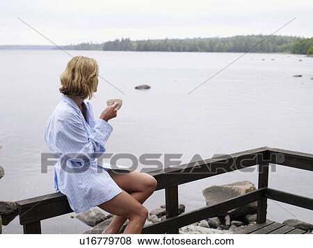 Stock Photo - Woman sitting on a porch by the water holding a mug.. Fotosearch