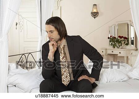 Stock Photography - young businessman sitting on bed holding chin in hand. Fotosearch