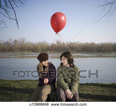 Boy (12-14) and girl (10-12) holding balloon, sitting by river, smiling View Large Photo Image Stock Photo - Boy (12-14) and girl (10-12) holding balloon, sitting by river, smiling. Fotosearch