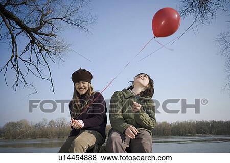 Boy (12-14) and girl (10-12) holding balloon, sitting by river, smiling View Large Photo Image Stock Photo - Boy (12-14) and girl (10-12) holding balloon, sitting by river, smiling. Fotosearch