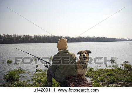 Stock Image - Boy (12-14) fishing by river with pet dog, rear view. Fotosearch
