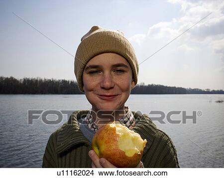 Boy (12-14) standing by river holding out apple, smiling, portrait View Large Photo Image Picture - Boy (12-14) standing by river holding out apple, smiling, portrait. Fotosearch