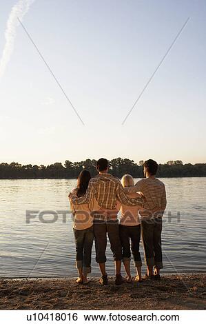 Stock Photograph - Four friends standing by a lake with arms around each other.. Fotosearch