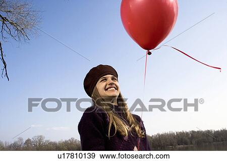 Stock Photo - Girl (10-12) outdoors, holding red balloon, smiling. Fotosearch
