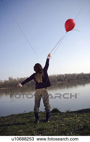 Stock Image - Girl (10-12) standing by river holding onto red balloon, rear view. Fotosearch