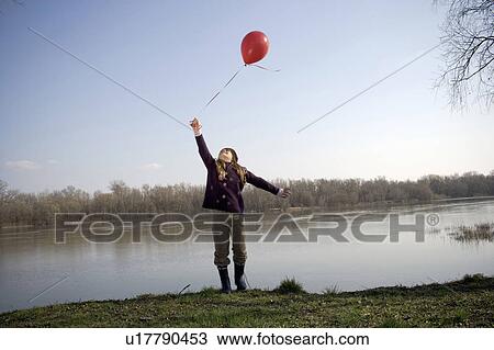 Stock Image - Girl (10-12) standing by river holding red balloon, looking upwards. Fotosearch