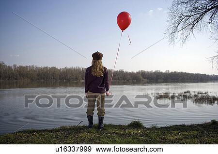 Stock Photo - Girl (10-12) standing by river holding red balloon, rear view. Fotosearch