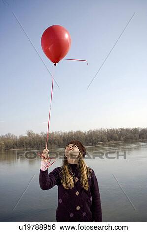 Stock Photograph - Girl (10-12) standing by river looking up at red balloon on ribbon. Fotosearch