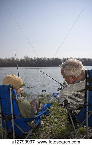 Stock Photo - Grandfather and grandson (12-14) fishing by river, rear view. Fotosearch