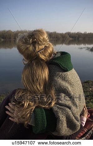Stock Photo - Grandmother and granddaughter (10-12) sitting by river, rear view. Fotosearch