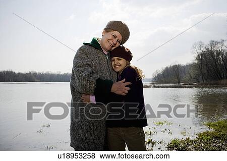 Stock Photo - Grandmother and granddaughter (10-12) embracing by river, smiling. Fotosearch