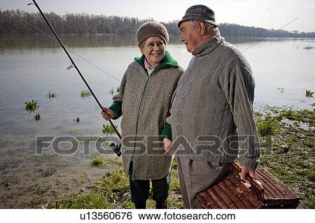 Stock Photograph - Senior couple carrying picnic basket and fishing rod by river, smiling. Fotosearch