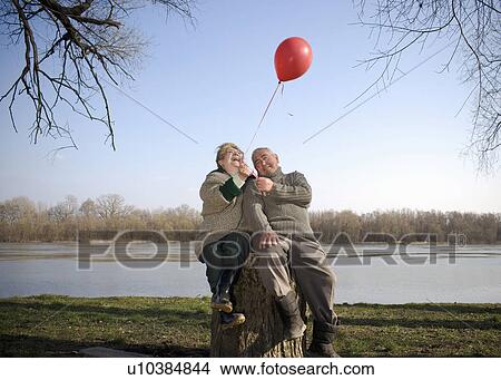 Senior couple sitting by river holding red balloon, smiling View Large Photo Image Picture - Senior couple sitting by river holding red balloon, smiling. Fotosearch