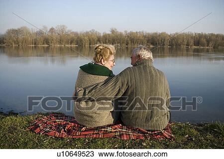Stock Image - Senior couple sitting on rug by river, rear view, woman smiling. Fotosearch