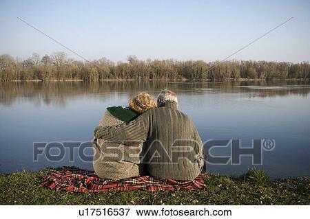 Stock Photo - Senior couple sitting on rug by river, rear view. Fotosearch