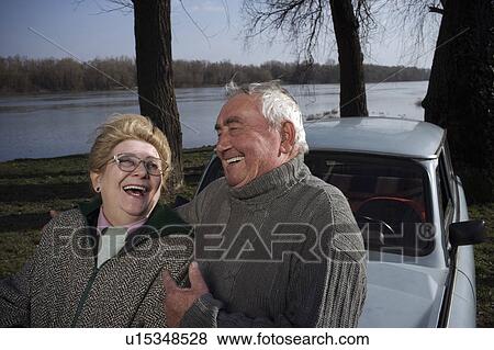 Stock Photo - Senior couple standing by car on riverside, laughing. Fotosearch