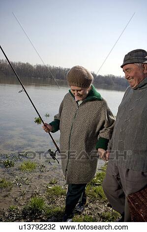 Stock Image - Senior couple walking by river, woman carrying fishing rod, smiling. Fotosearch