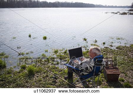 Stock Photo - Senior man fishing by river, using laptop, rear view. Fotosearch