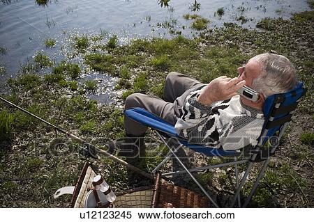Stock Photography - Senior man fishing by river, using mobile phone, overhead view. Fotosearch