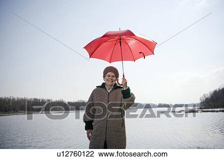Stock Image - Senior woman standing by river holding red umbrella, smiling, portrait. Fotosearch