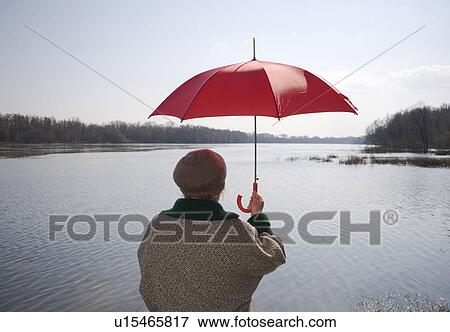 Stock Photo - Senior woman standing by river holding red umbrella, rear view. Fotosearch
