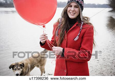 Stock Photograph - Young woman holding balloon walking dog by river, smiling, portrait. Fotosearch