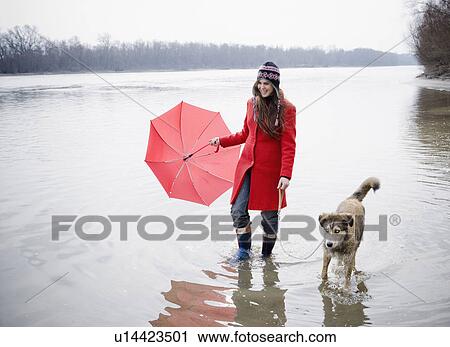 Stock Image - Young woman holding umbrella, wading in river with dog, smiling. Fotosearch