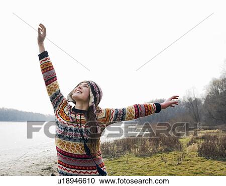Stock Image - Young woman standing by river with arms outstretched, smiling. Fotosearch