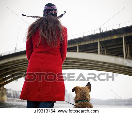 Picture - Young woman wearing hat standing by river with dog, rear view. Fotosearch