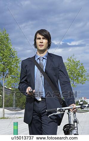 Stock Photo - Businessman in park walking his bike with headset and mobile frontal view.. Fotosearch