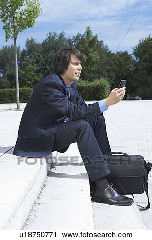 Stock Image - Businessman sitting on steps looking at his mobile view from side park in background.. Fotosearch