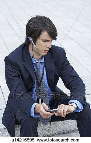Stock Photography - Businessman sitting on steps looking at his watch with headset and mobile.. Fotosearch