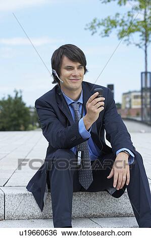 Stock Photograph - Businessman sitting on steps with headset looking at his mobile.. Fotosearch