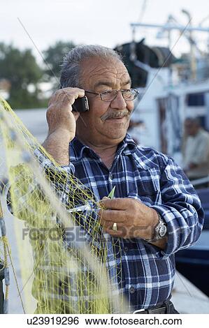 Stock Photograph - Mature fisherman fixing net while on cell phone.. Fotosearch