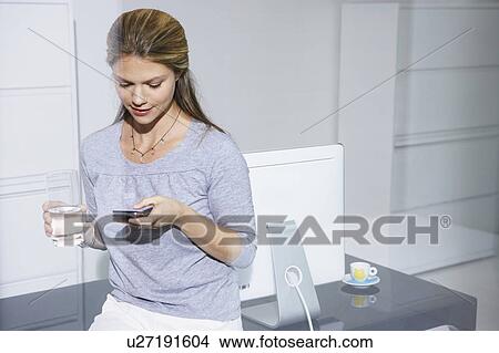 Woman sitting on computer desk looking at cell phone. View Large Photo Image Picture - Woman sitting on computer desk looking at cell phone.. Fotosearch